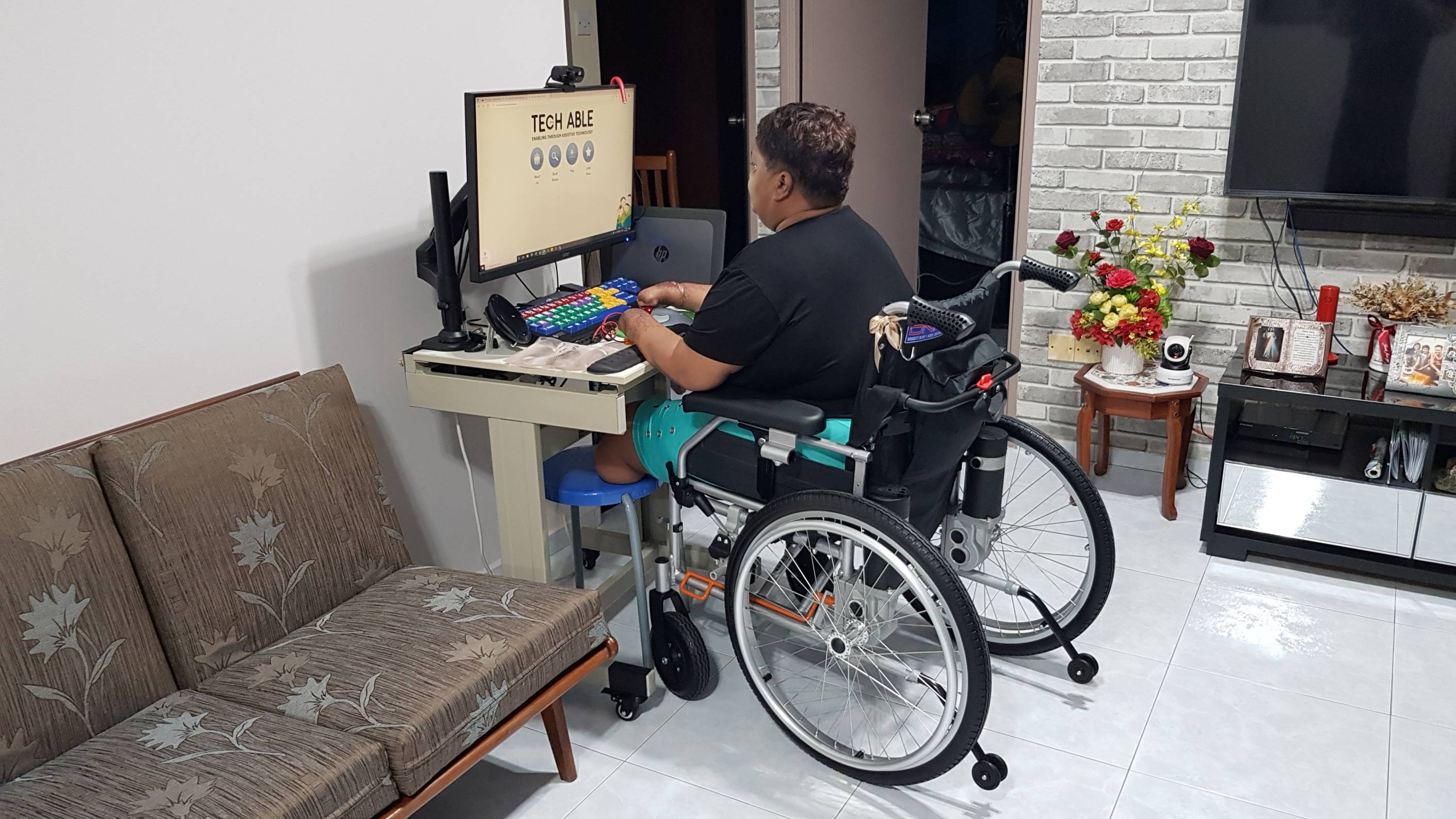 Carole sitting in a wheelchair and looking at her computer monitor screen placed on top of a height-adjustable desk. The desk also contains some of the other assistive technology devices she uses for doing her work. She places her right residual limb on the trackball of an adapted mouse, while her left residual limb is near a 3D-printed assistive switch in orange, which is connected to the trackball mouse’s left button.